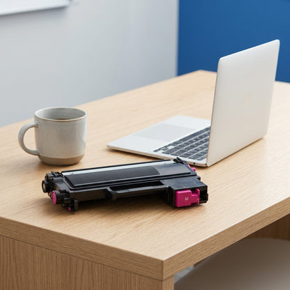 Brother TN225M magenta toner cartridge shown for scale next to a laptop and mug on an office desk.