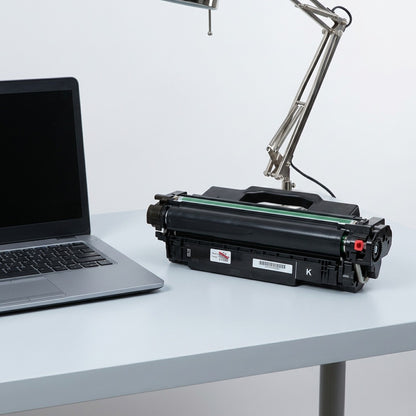 HP 507X CE400X black toner cartridge on a desk next to a laptop and lamp for scale reference.