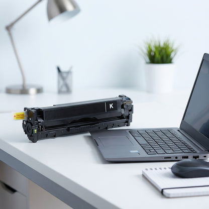 A black HP 131X (CF210X) toner cartridge next to a 14-inch laptop on a white desk for scale.