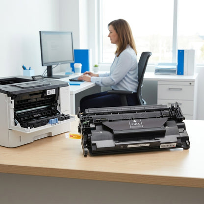 HP 26X CF226X toner cartridge on an office desk next to a printer with an employee in the background.