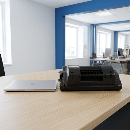 HP 81X (CF281X) compatible black toner cartridge on an office desk next to a laptop for scale.