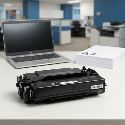 High-yield black toner cartridge for HP 87X on a desk next to a laptop and A4 paper for scale.