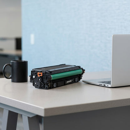 HP 508A (CF360A) black toner cartridge on an office desk next to a laptop and coffee mug for scale.