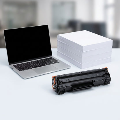 A black Elevate Imaging HP 85A toner cartridge on a desk beside a laptop and a ream of paper.